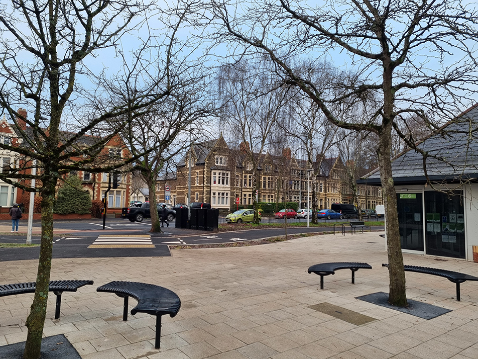 A photograph showing public realm improvements at the entry to the Penylan Community Centre and Library, including new seating, trees and cycle parking in the foreground, and in the background showing bi-directional cycle track and pedestrian zebra crossing.