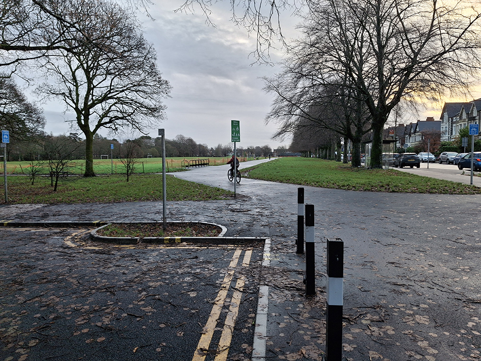 A photograph showing a shared use space in the foreground at the western closed off end of Alder Road, along with bollards and a “share with care”, with a cyclist on the shared space in the background entering the cycle track.