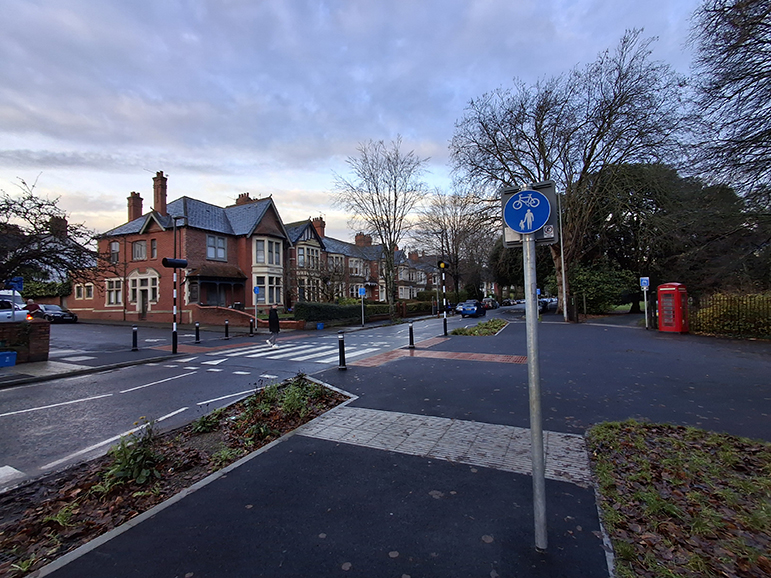 A photograph showing a shared use space and rain garden in the foreground and at the background a pedestrian on a zebra crossing, tabled/ramped with parallel cycle crossing, at Ninan Road, with modal filter to Kelvin Road.