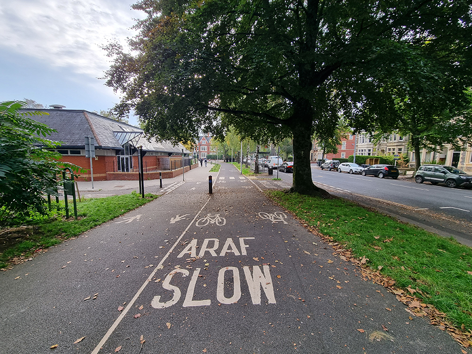 A photograph showing widened footpath next to a bi-directional cycle track in the Roath Recreational Grounds, along Ninan Road in the foreground, and a pedestrian zebra crossing over a bi-directional cycle track in the background.