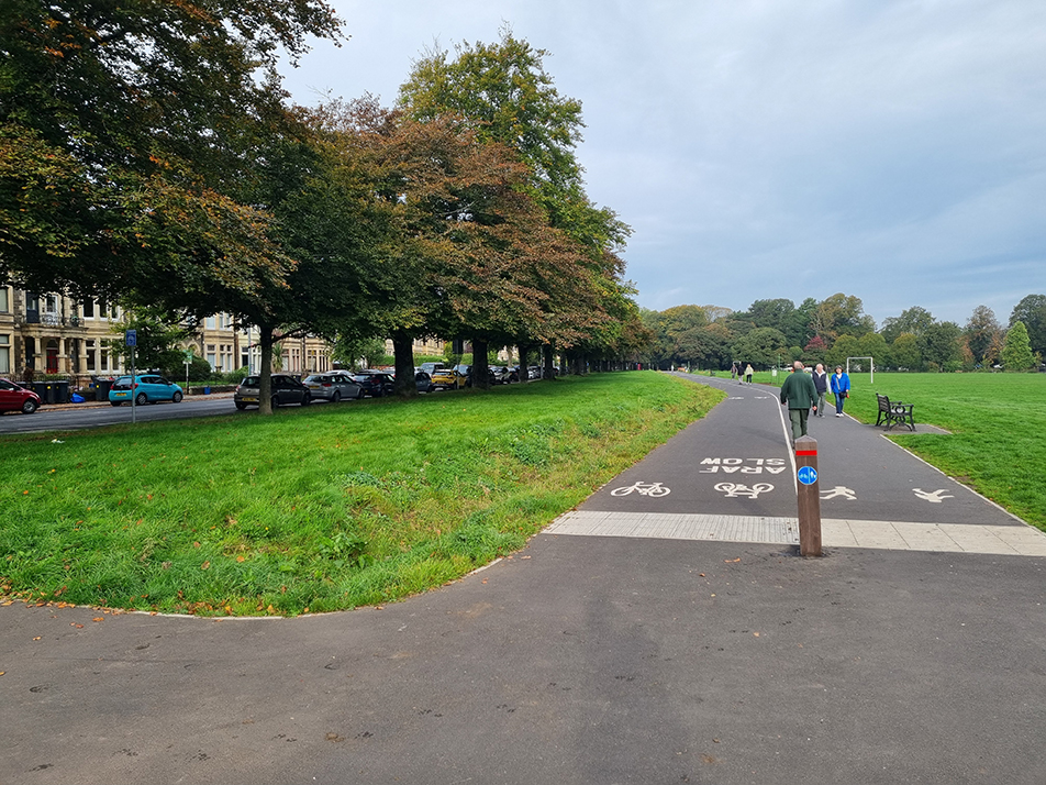 A photograph showing widened footpath and the bi-directional cycle track with pedestrians in the background, and a shared use space in the foreground, at the Roath Recreational Grounds, which links into the zebra crossing on Ninan Road near Pen-Y-Wain Place.