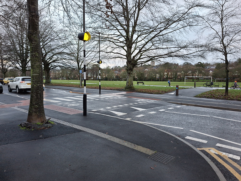 A photograph showing a zebra crossing tabled/ramped with parallel cycle crossing, at Ninan Road, near Pen-Y-Wain Place, in the foreground and the link to the Roath Recreational Grounds cycle track.