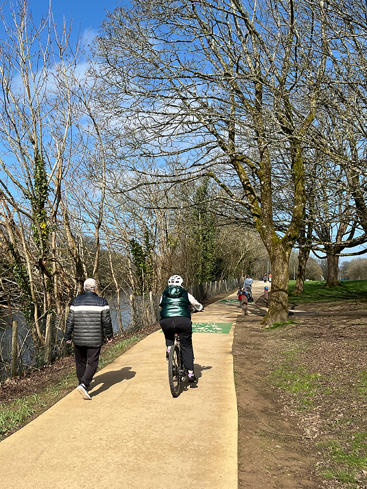 A cyclist cycling on a buff coloured path through a park on a sunny day with a person walking alongside them.  Green share with Care path marking visible on the path surface.  Person pushing a pushchair walking towards the camera. Path is tree lined.