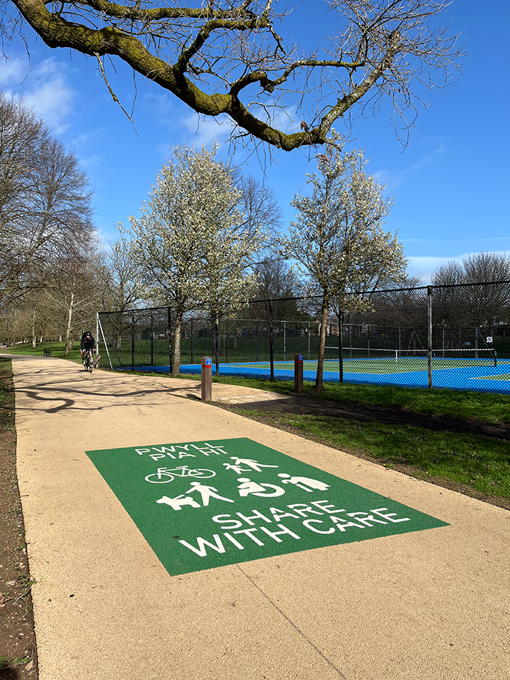 Male cyclist cycling on a buff coloured path through a park on a sunny day adjacent to a tennis court. Green share with Care path marking visible on the path surface